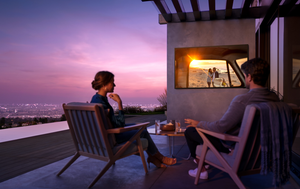 Couple sitting on patio overlooking city in background watching a weather-proof TV mounted on the wall at sunset. The TV is showing a movie with a couple on the beach behind a car window. Writing on picture says Relax, work, and play outdoors. Tech helps 