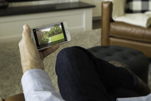 Mans hand holding an iPhone displaying the smart home surveillance app showing the view of the backyard. 