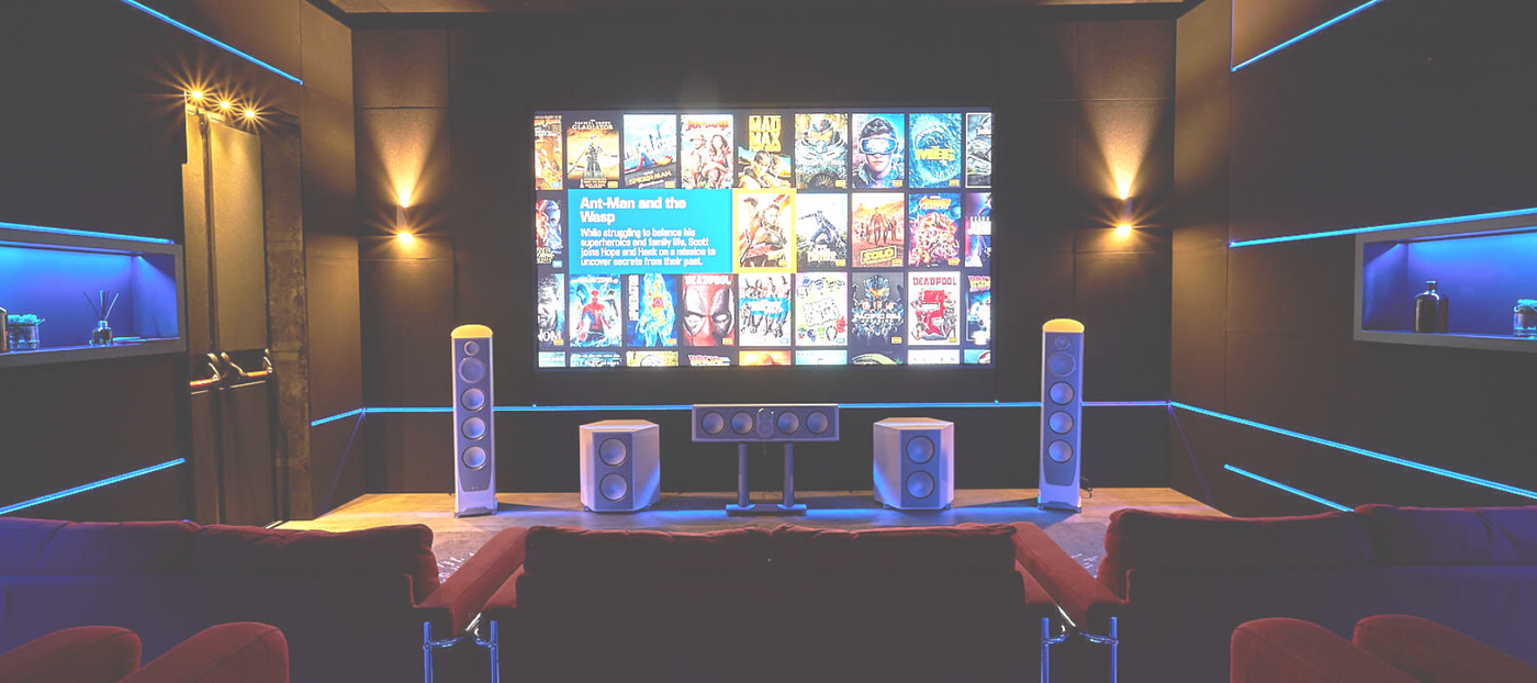 Home theater room with large screen showing movie selections on it. Five free standing white paradigm speakers place under screen. Red theater seating in background. Text on image says Feel the music as it was intended. 