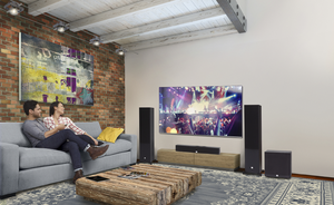 Couple sitting on a couch in the family room watching a large TV mounted on the wall. Free standing speakers are placed on the floor around the TV. 