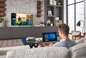 Man sitting on a couch in living room working on a laptop computer. Built-in gray entertainment center with large TV sitting on top. TV shows the back of a woman in a kayak in the ocean surrounded by mountains at sunset. 