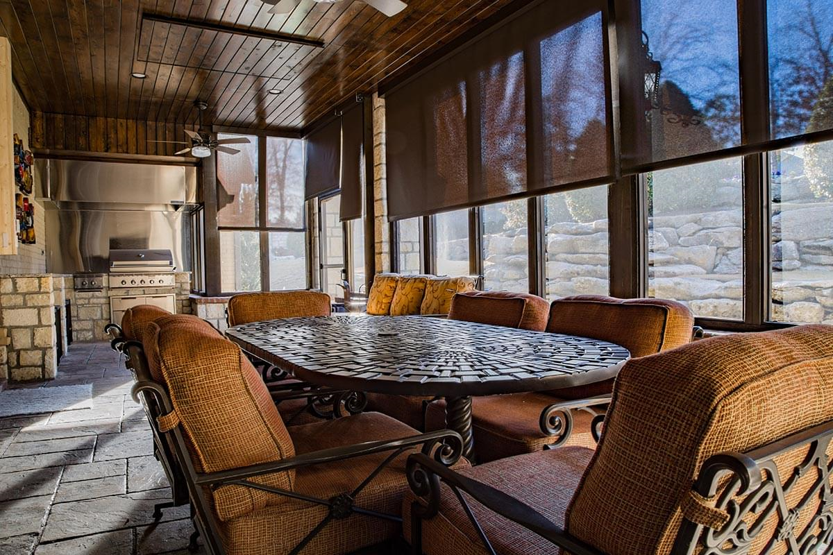 Large enclosed outdoor patio with raw iron table and cushioned chairs. The windows are covered by brown motorized shades. There is a built-in grill and kitchen. 